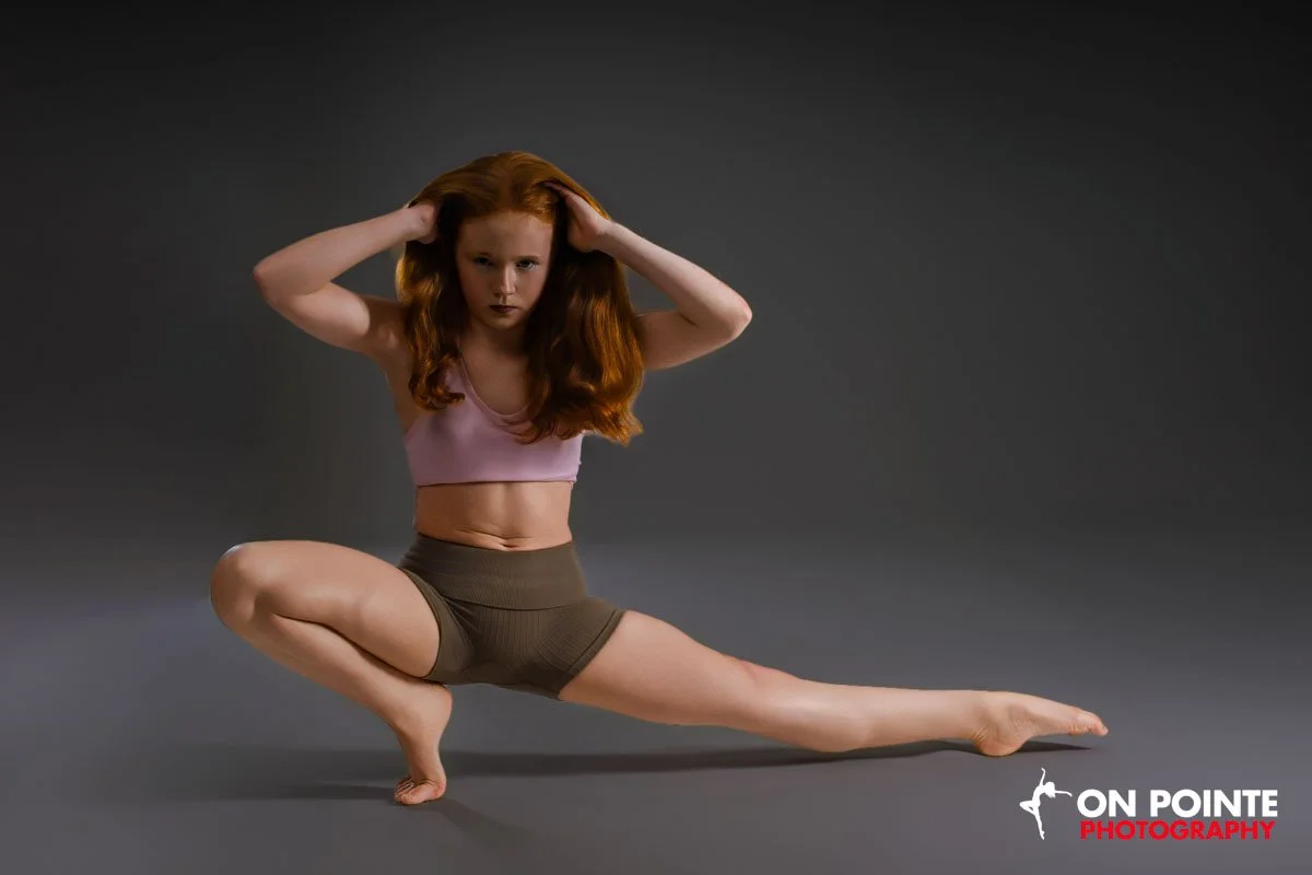 A woman with long red hair performing a yoga pose on a gray studio background. She is wearing a pink sports bra and brown shorts, balancing with one hand on the ground and her other hand on her head.