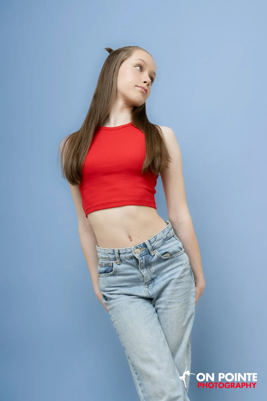 Woman in red crop top and jeans posing against blue background.