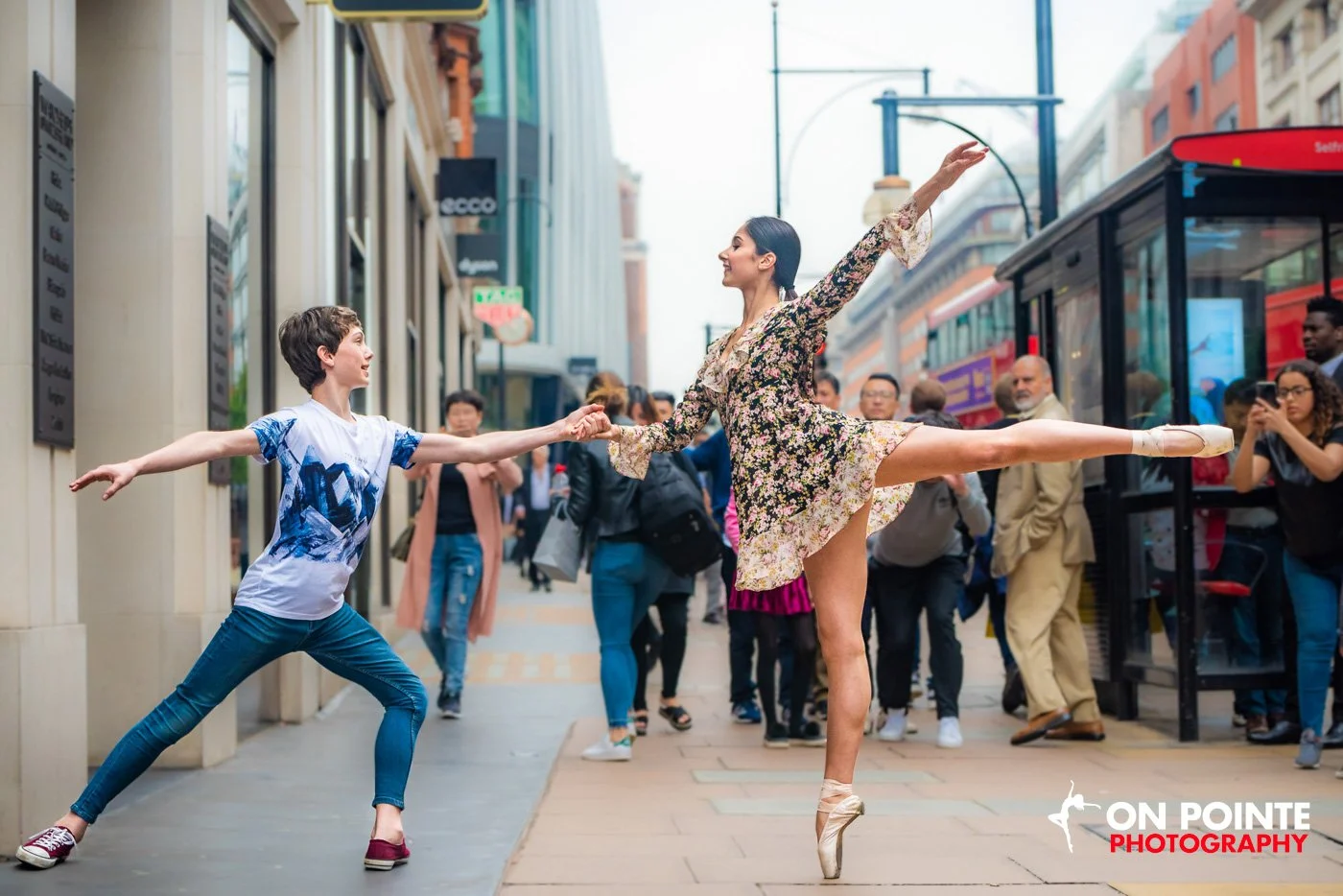 Two ballet dancers perform on a busy city street, one in a floral dress en pointe and the other in casual attire, surrounded by onlookers and buses.