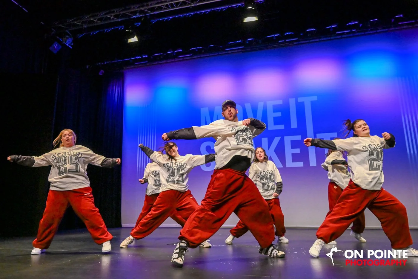 A dance performance on stage featuring six dancers wearing gray jerseys with black sleeves and red baggy pants, striking energetic poses. The stage has a blue and purple lit background with the words 'MOVE IT SOCKER' visible, and a logo 'ON POINTE PH