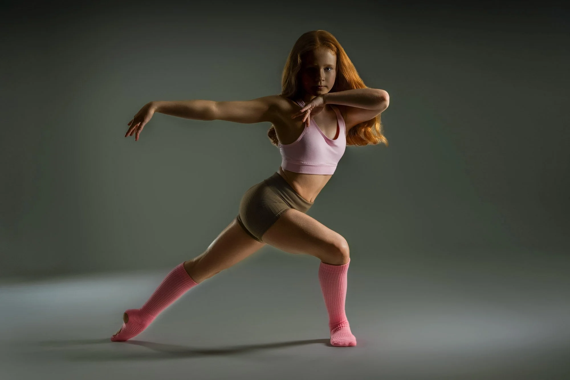 A young woman with red hair in athletic wear, stretching in a lunge pose with one arm extended and the other bent, on a plain gray background.