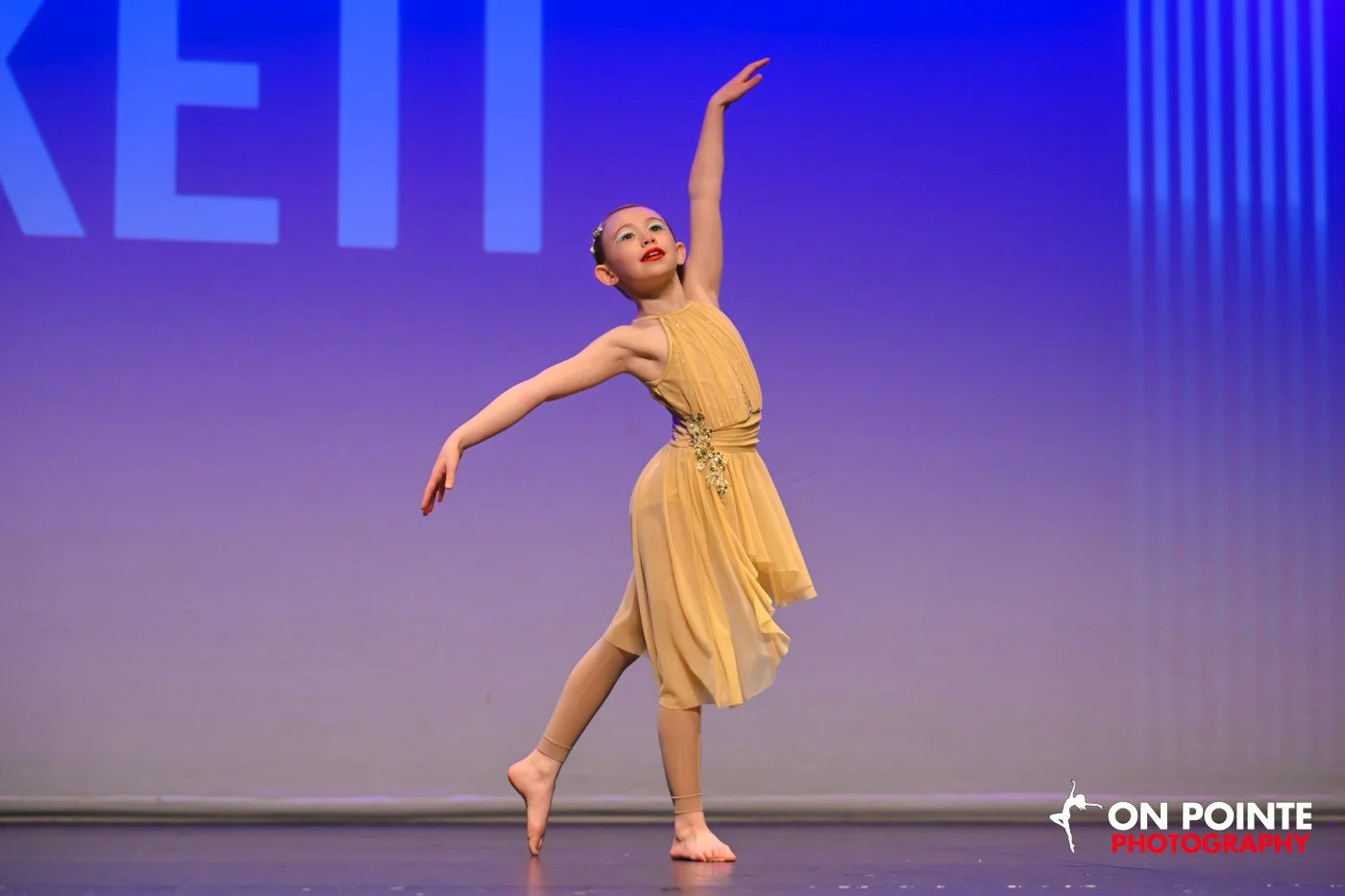 Young girl ballet dancer in a yellow dress performing a dance on stage with her arms raised, on a purple background.