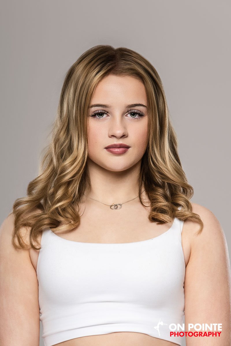 Young woman with long, wavy blonde hair wearing a white tank top and a delicate necklace, looking directly at the camera against a neutral background.