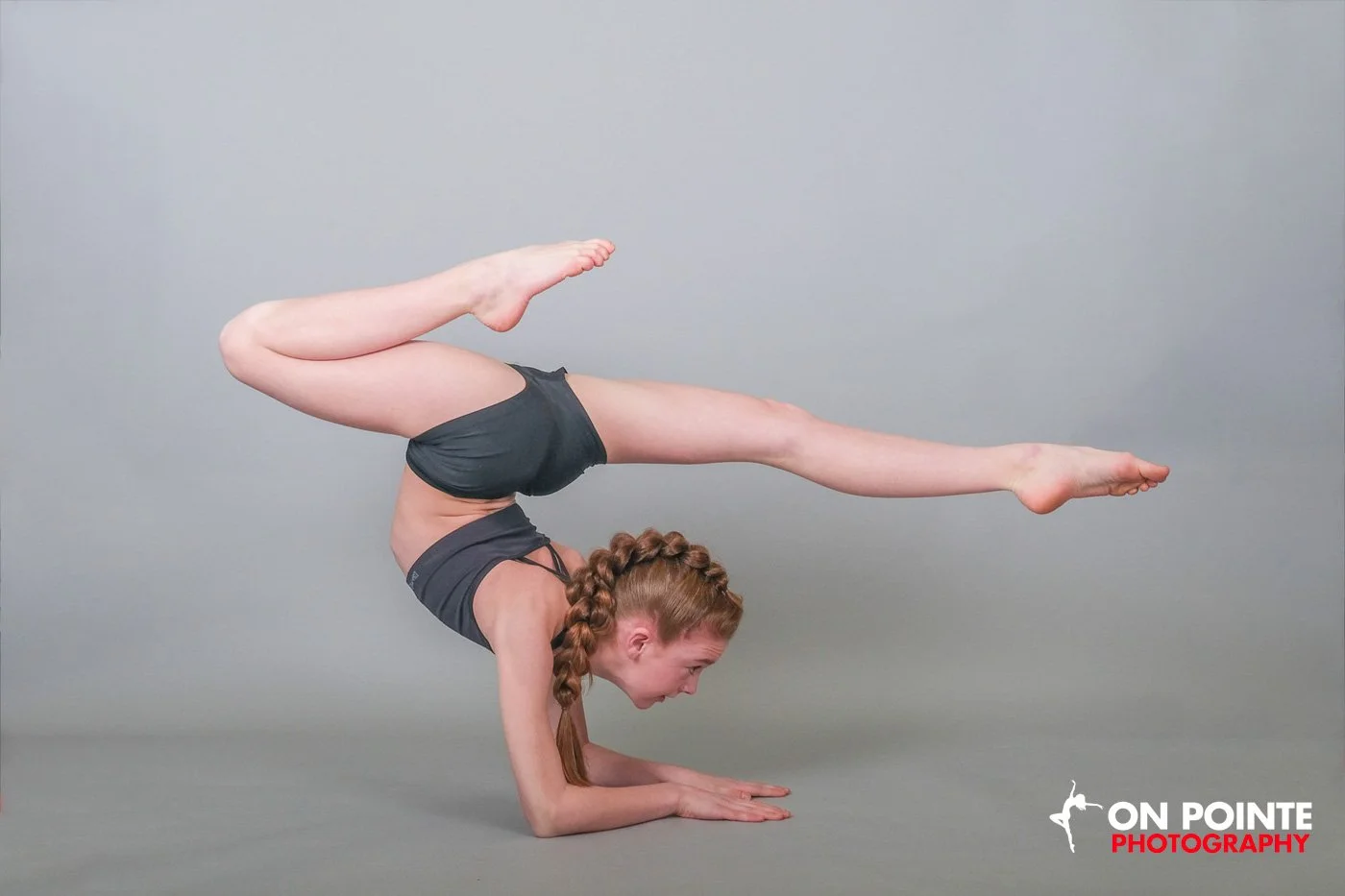 A girl performing a complex yoga pose on a gray background. She is balancing on her forearms with her legs extended backward and upward. She has braided hair and is wearing dark athletic wear.