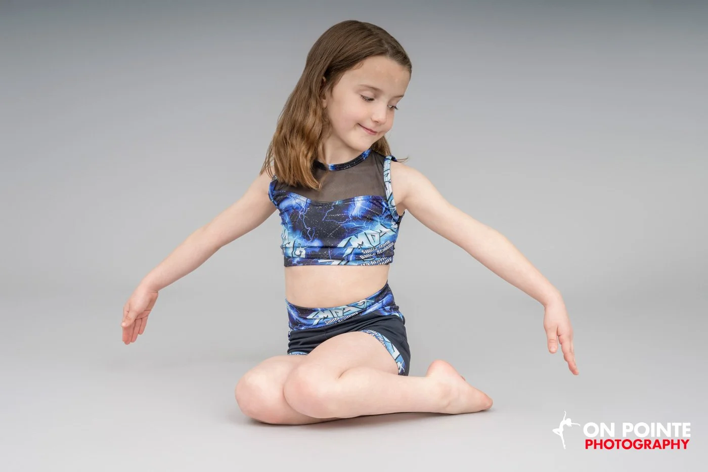 A young girl in a blue dance outfit poses gracefully on her knees with her arms extended. She is performing a dance move in a studio setting.