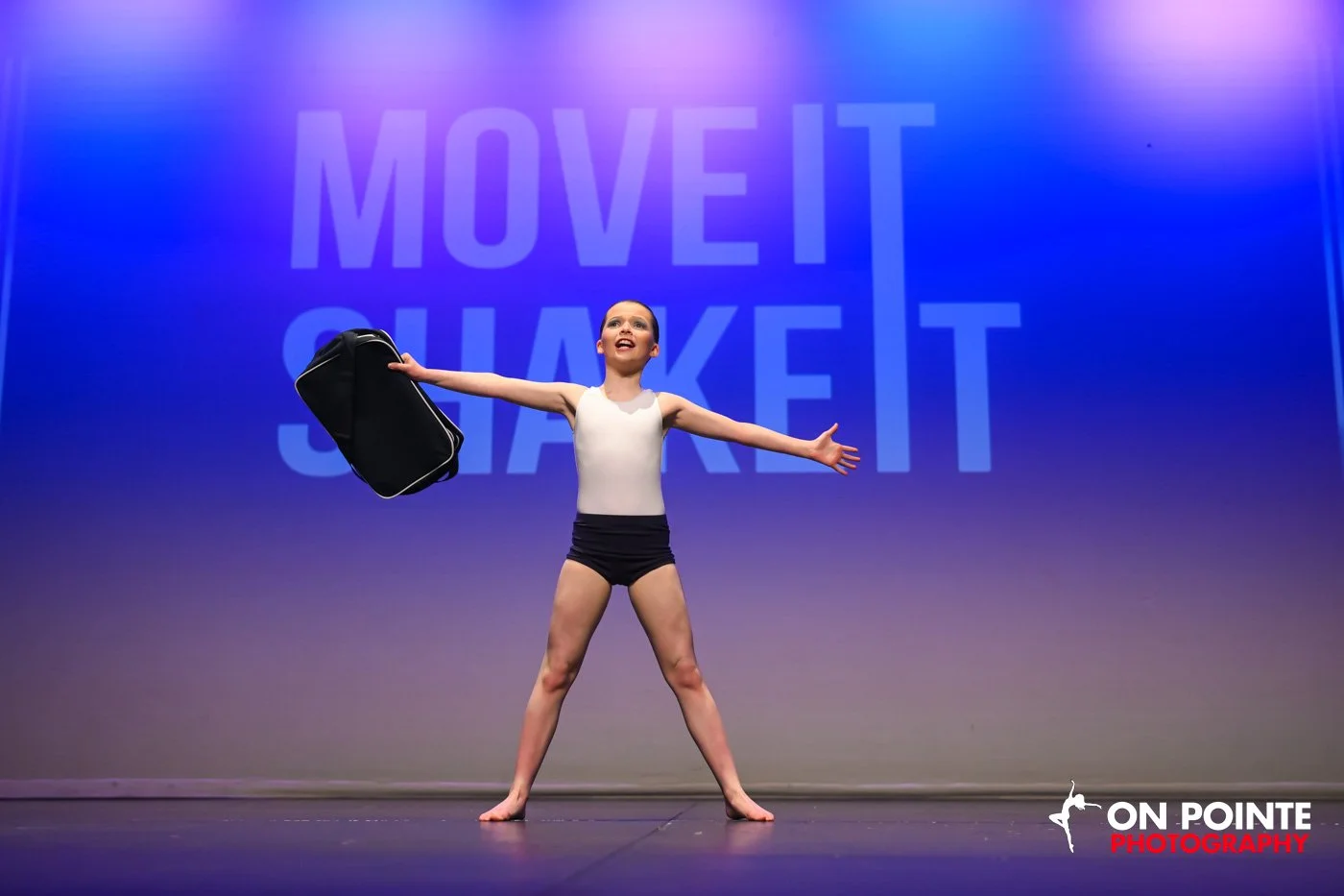 A young girl in dance attire on stage, holding a black bag with one hand, with arms outstretched and a joyful expression, performing in front of a large screen displaying the text 'MOVE IT QUIET'
