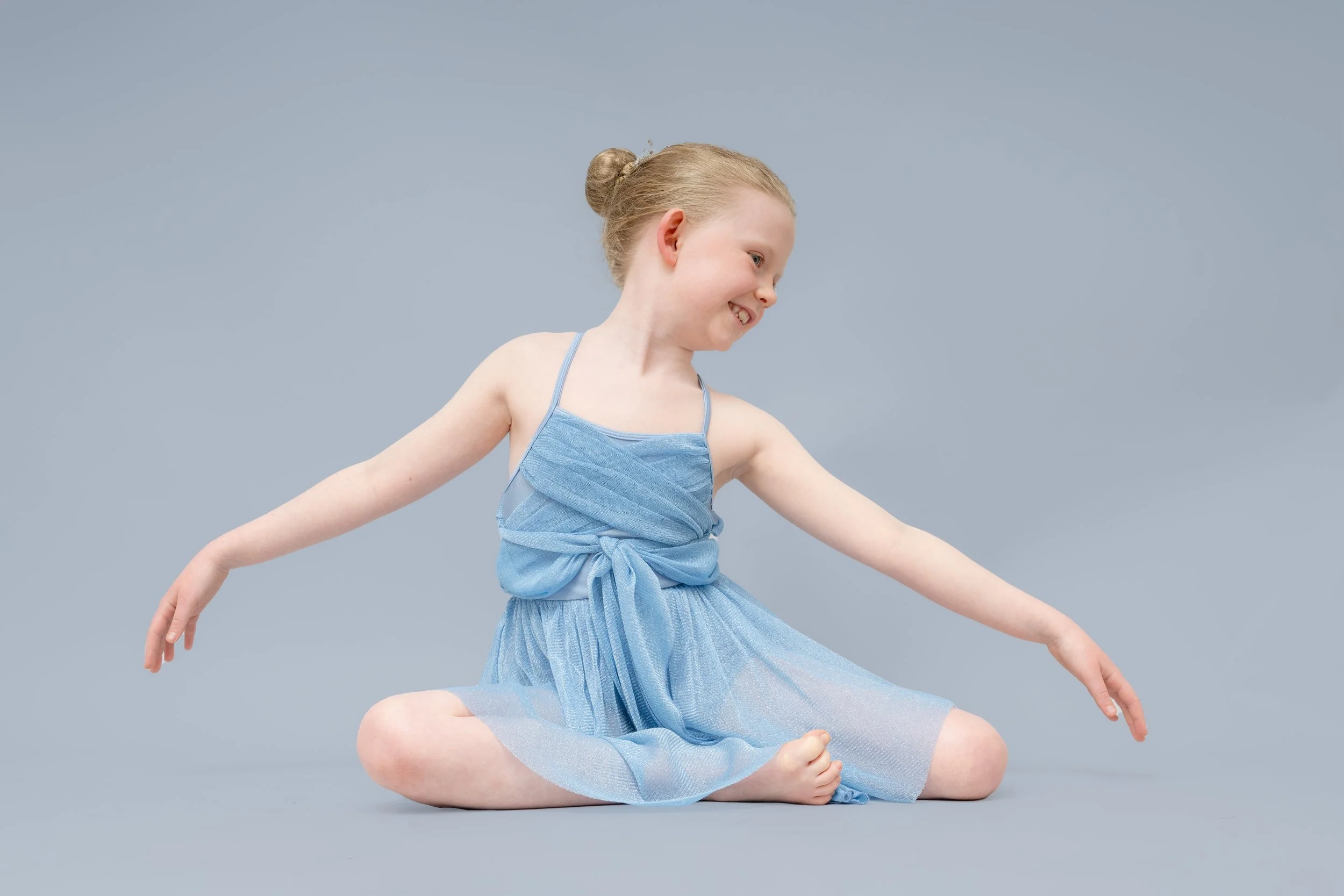 Young girl in a blue dress sitting on the floor with arms and legs extended, smiling and looking to the side against a plain background.