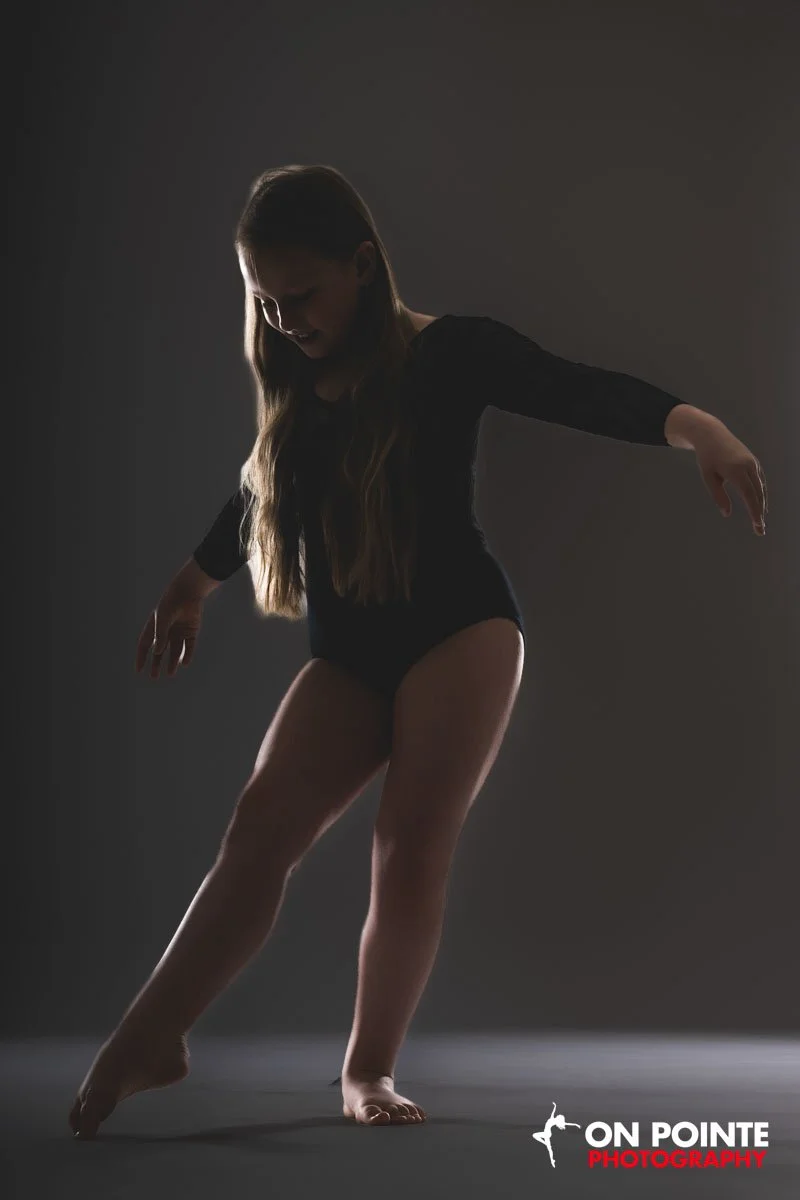 A young woman with long hair wearing a black leotard or ballet outfit, striking a dance pose with one leg extended and arms outstretched, against a dark background. The photo is a professional dance performance shot.