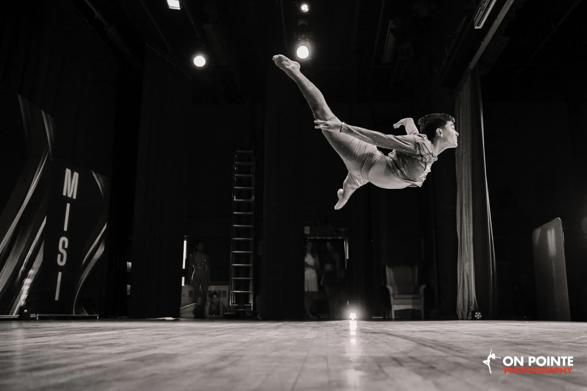 A gymnast performing an arabesque on a stage with a dark background, overhead lights, and a ladder in the background, with the logo 'ON POINTE PHOTOGRAPHY' in the bottom right corner.