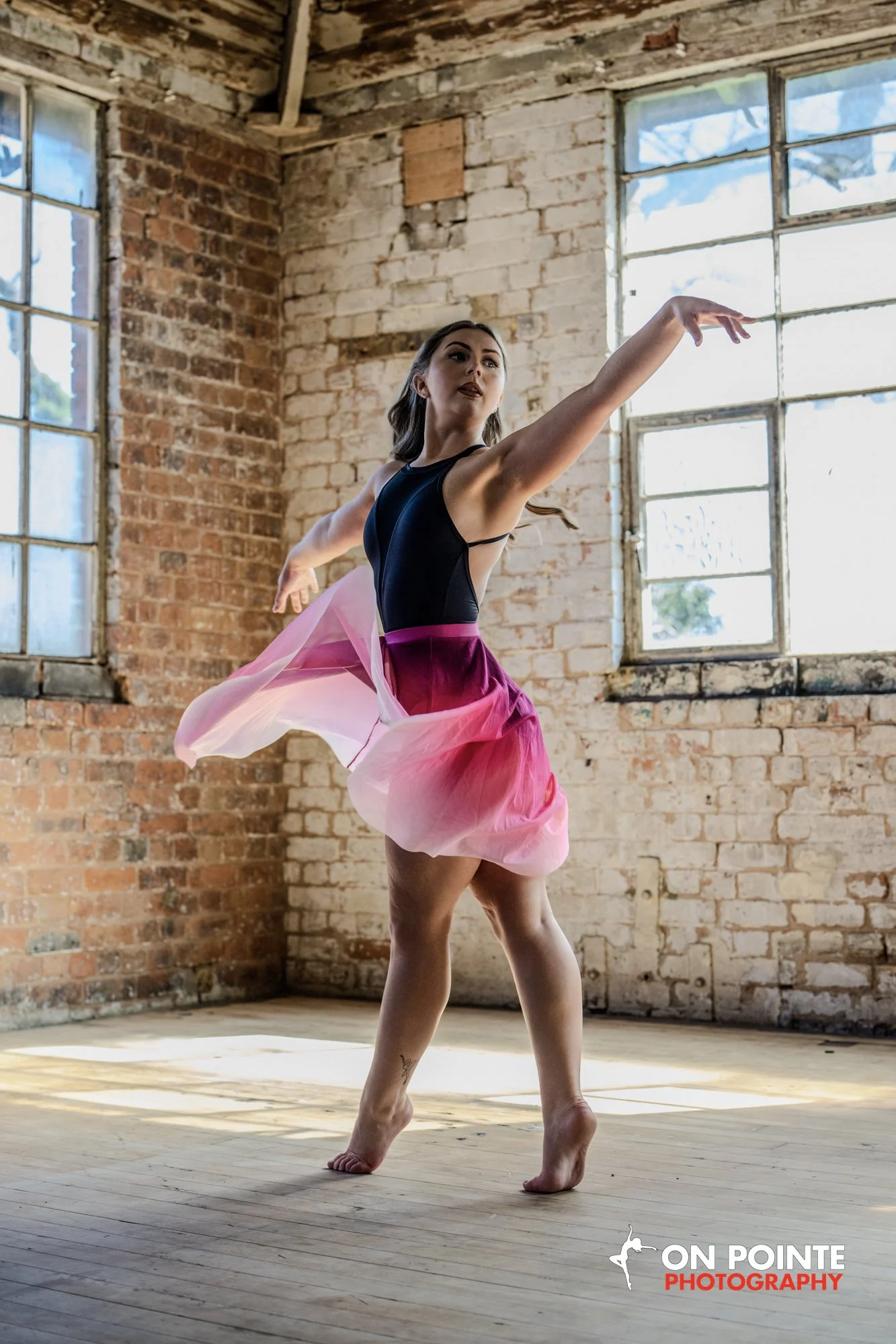 A ballerina practicing in an industrial-style room with exposed brick walls and large windows, wearing a black top and a pink flowing skirt.