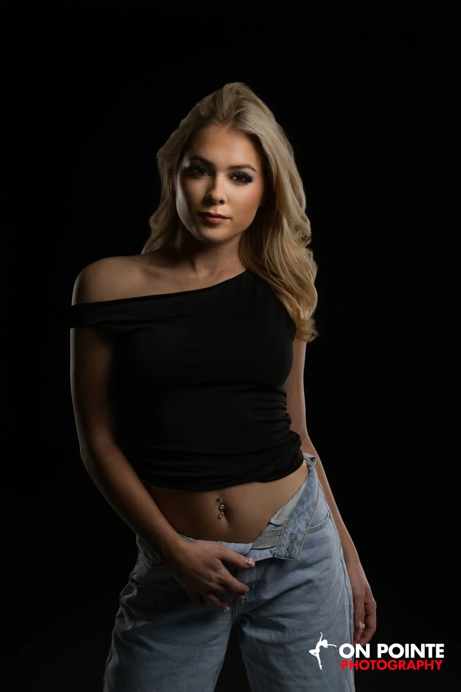 Woman with long blond hair wearing an off-shoulder black top and jeans, posing against a black background.