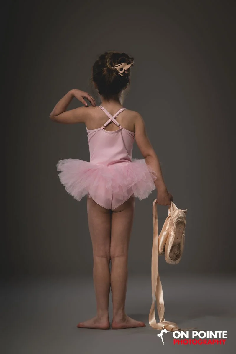 A young girl wearing a pink ballet tutu, holding ballet shoes, and wearing a mask, standing against a plain gray background.