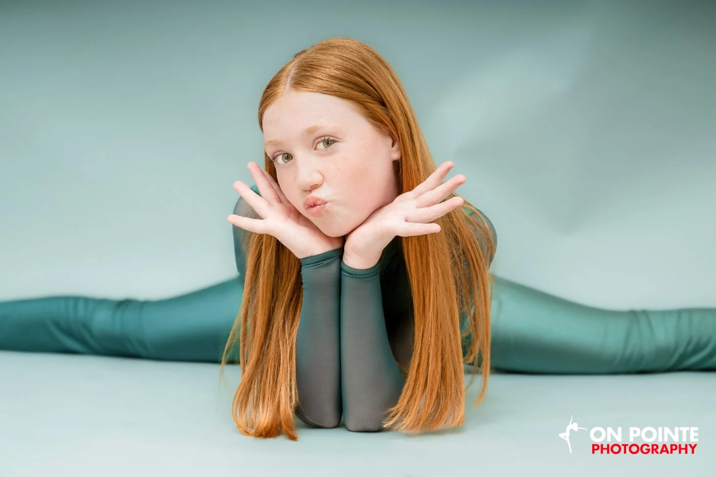 Young girl with long red hair in a split pose, wearing a teal outfit, hands under chin, smiling playfully, light green background.
