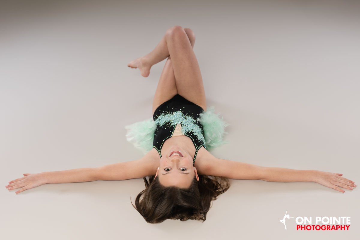 A young woman lying on the floor with arms and legs spread out, wearing a black leotard with light blue embellishments and a fluffy green skirt.