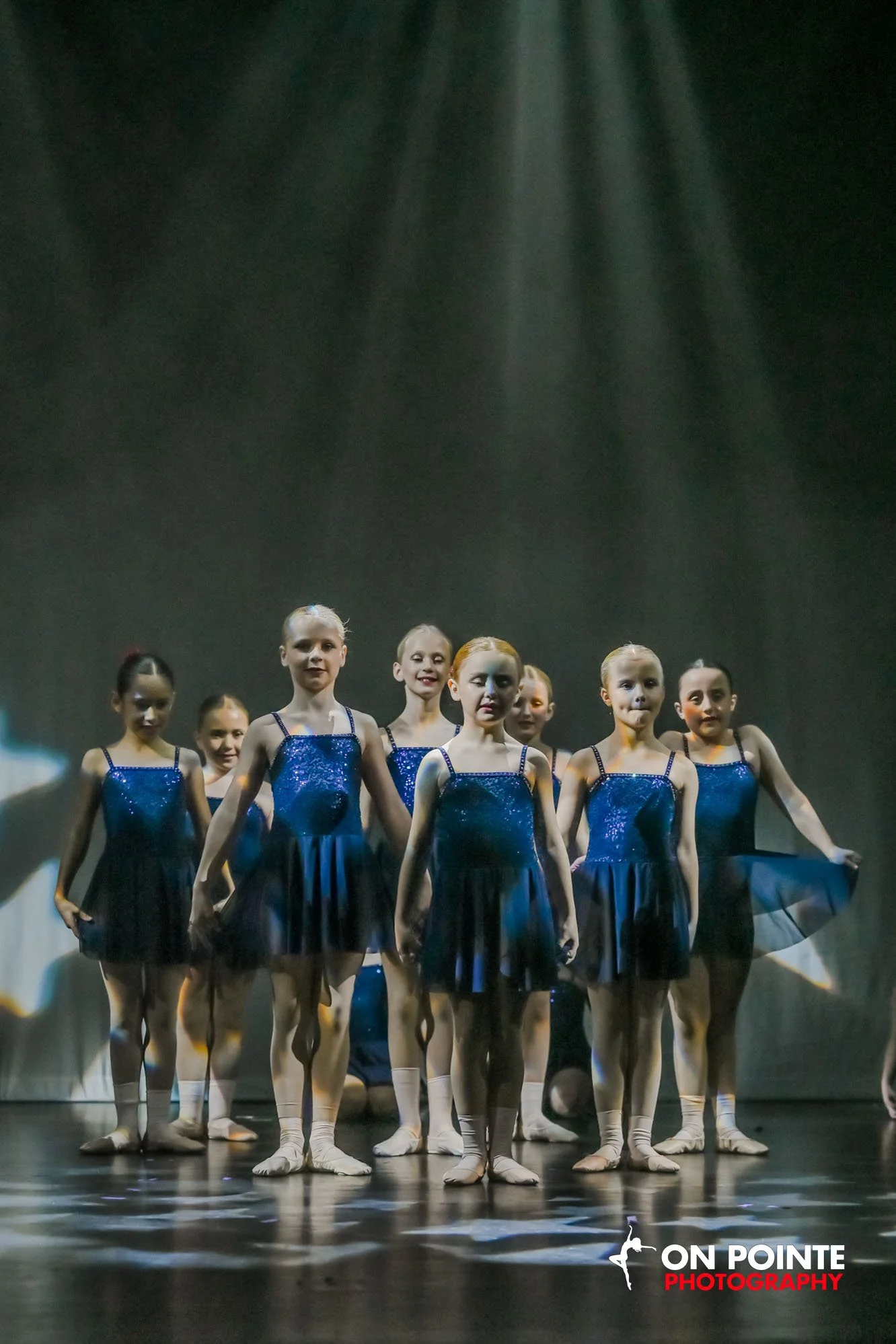 A group of young ballet dancers standing on stage in a formation, wearing blue costumes and beige tights, with stage lighting above.