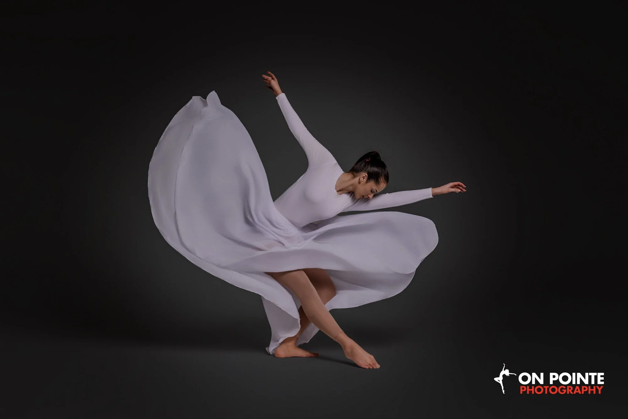 A female dancer in a white dress mid-spin against a dark background, with her arms extended and one leg bent.