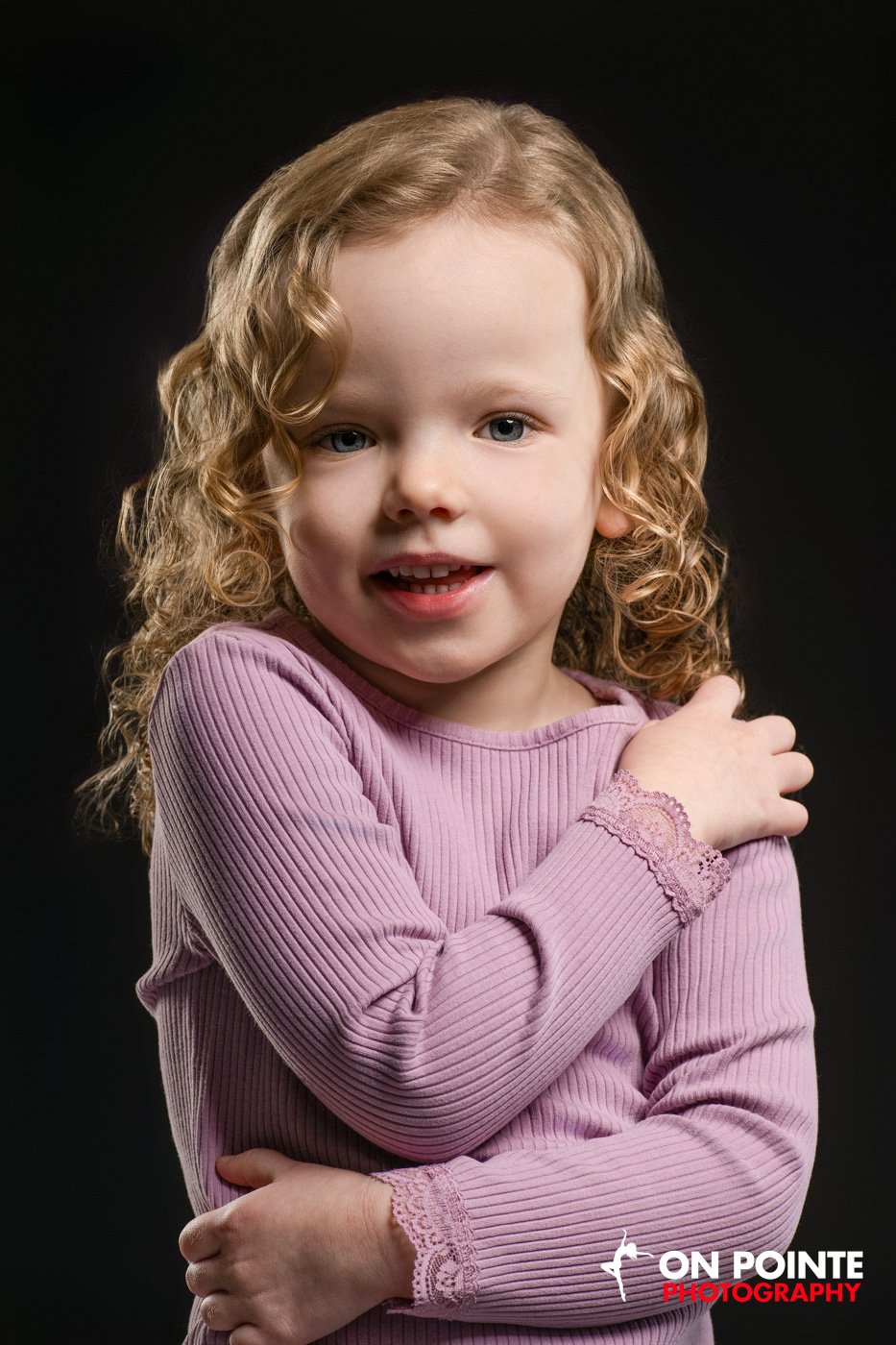 Young girl with curly hair wearing a pink sweater, posing against a dark background.