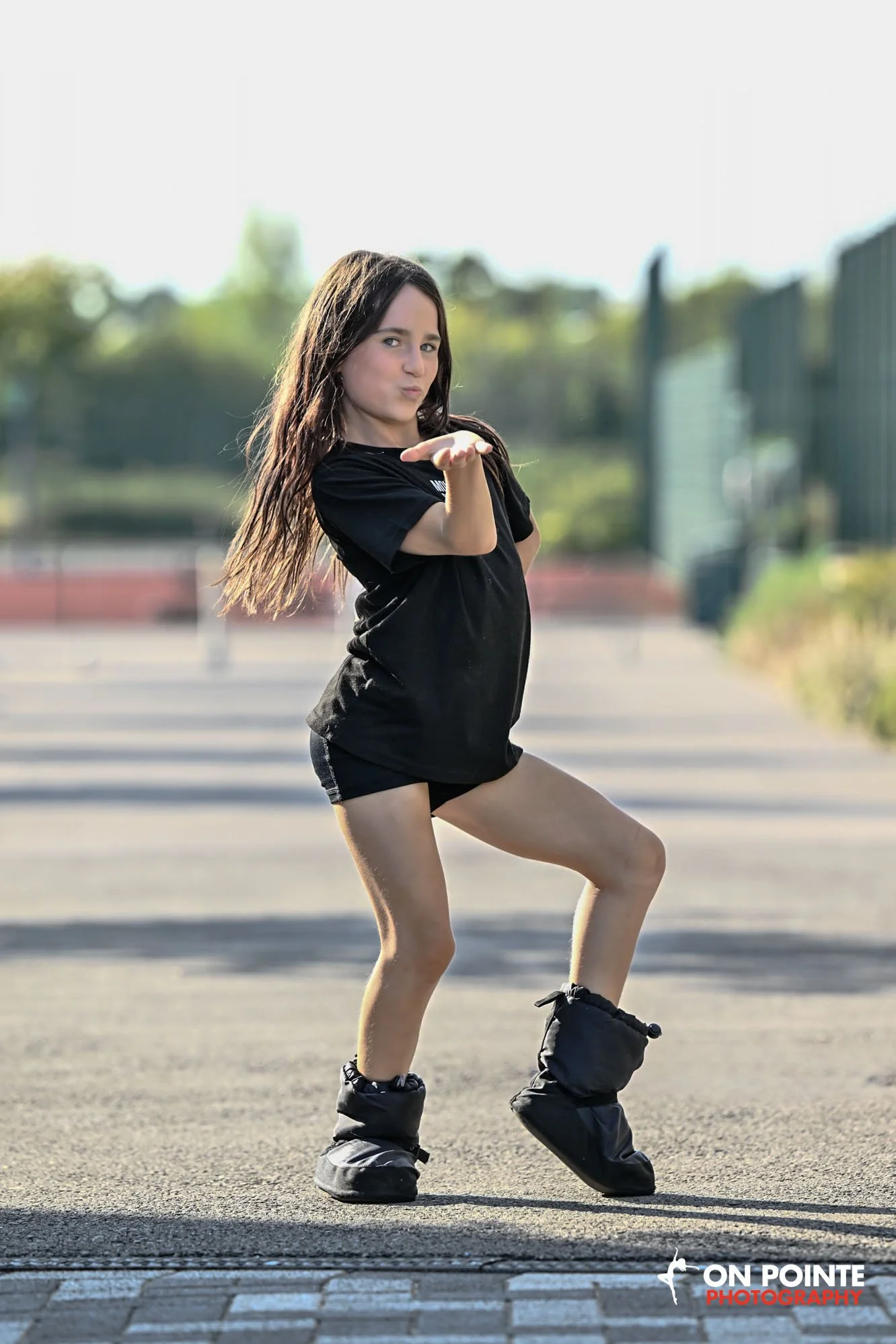 Young woman in black dance attire performing on an outdoor paved area with barriers and greenery in the background, blowing a kiss to the camera.