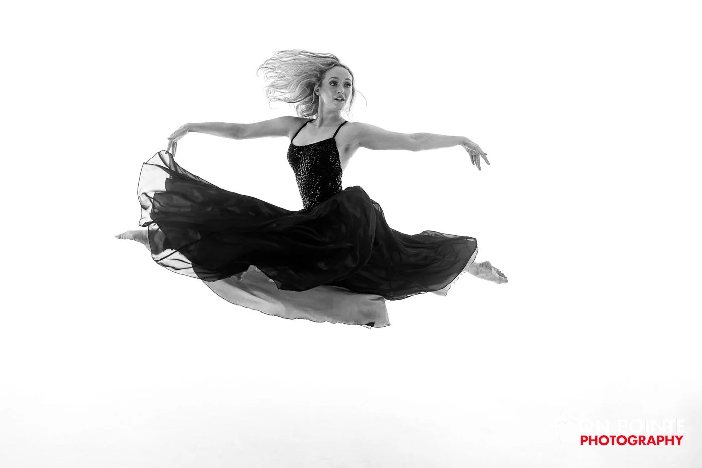 Ballet dancer performing a grand jeté in mid-air, wearing a flowing black dress, on a white background.