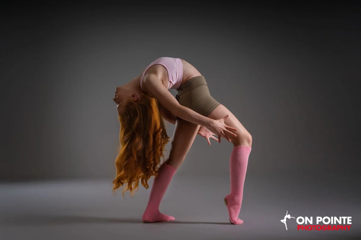 A woman with long, curly red hair is performing a contorted dance pose in a studio with a dark background. She is wearing a pink top, beige shorts, and pink knee-high socks.