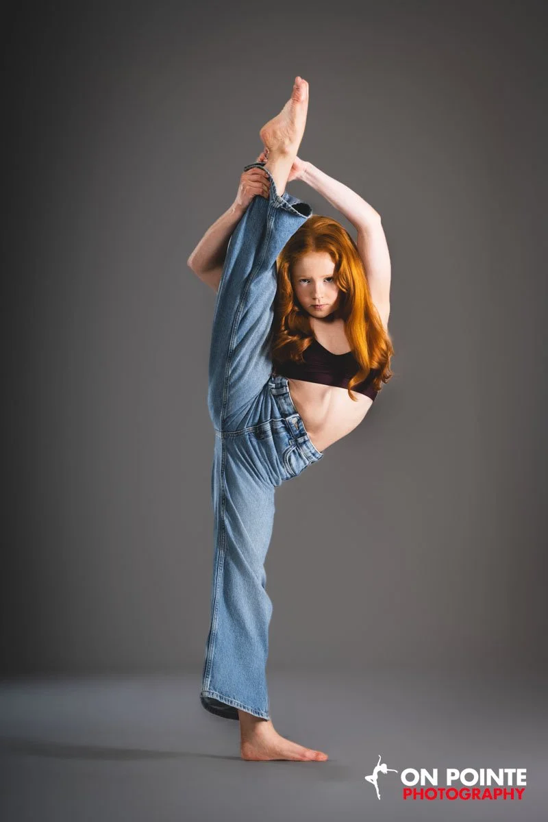 A young woman with long red hair practicing a high flexibility yoga pose on a gray backdrop, balancing on her left foot with her right leg lifted and held upward, wearing blue jeans and a black sports bra.