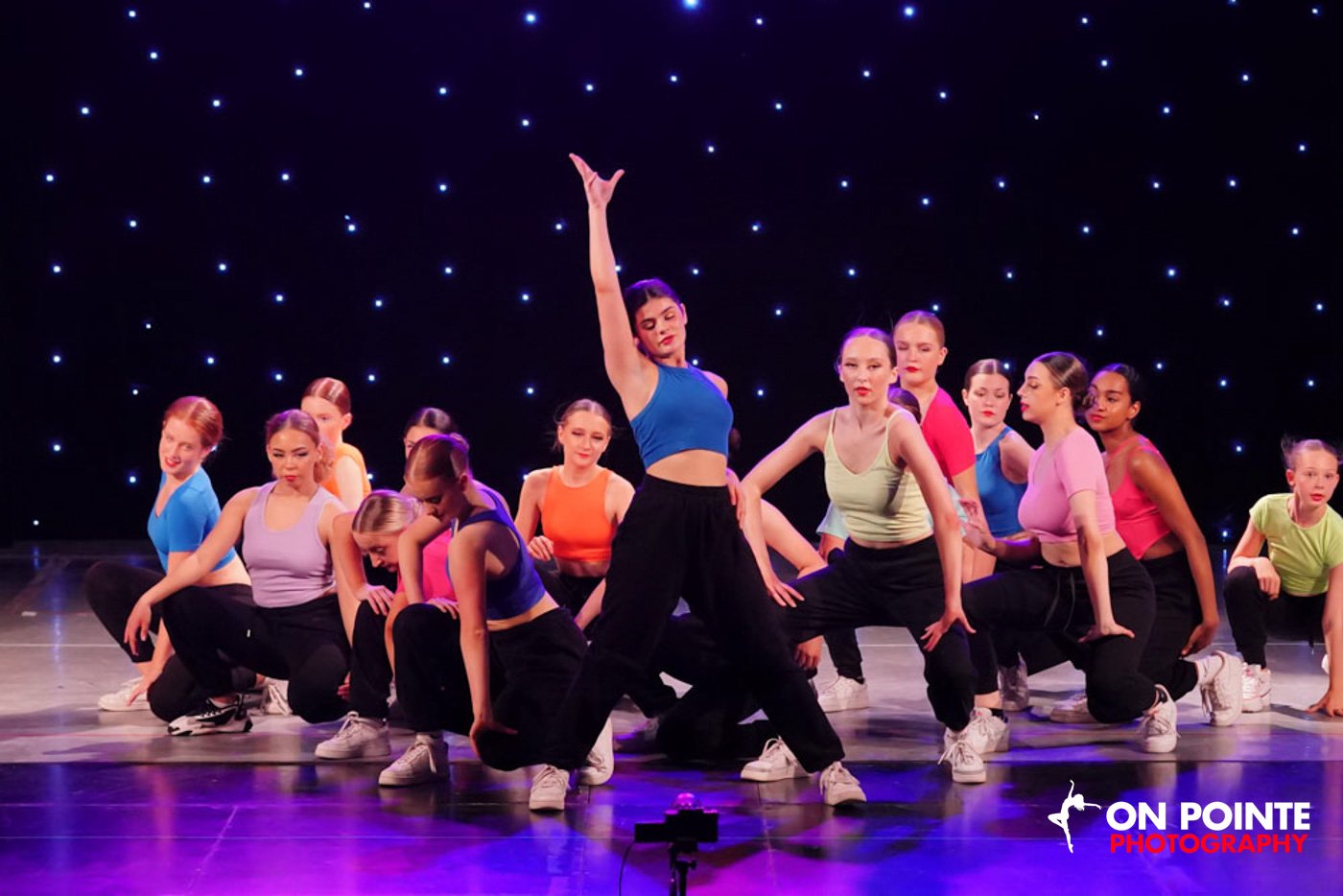 Group of young female dancers performing on stage with a starry backdrop. One dancer in the center strikes a pose with one arm raised, wearing a blue crop top and black pants. All dancers wear colorful tops and black pants.
