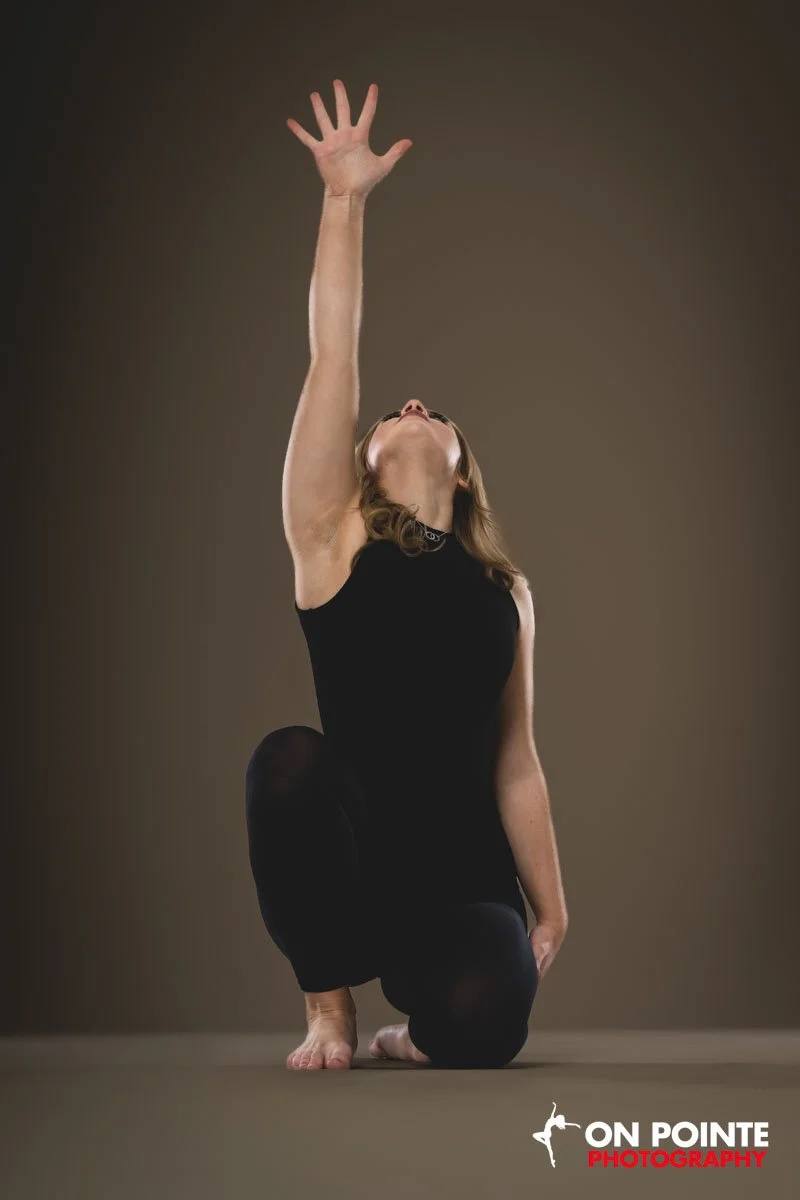 A woman in black clothes is kneeling on one knee with her other leg bent, reaching upward with her right arm and hand extended towards the sky, in a yoga or dance pose, against a plain background.