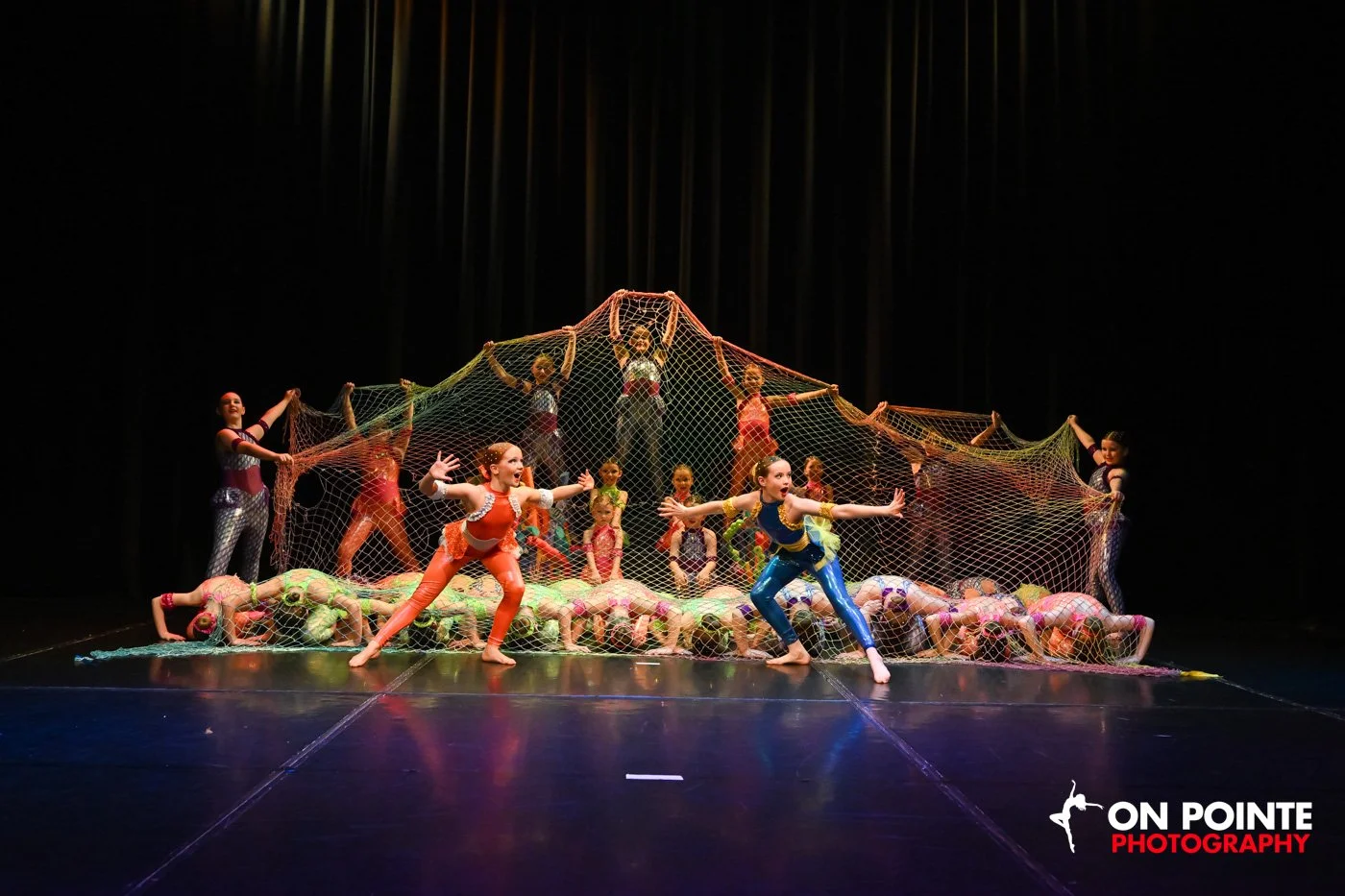 Dance performance on stage with children and teenagers interacting with a large colorful net, with some children crawling underneath and others holding the net, with a dark curtain background and a logo 'On Pointe Photography' in the bottom right cor