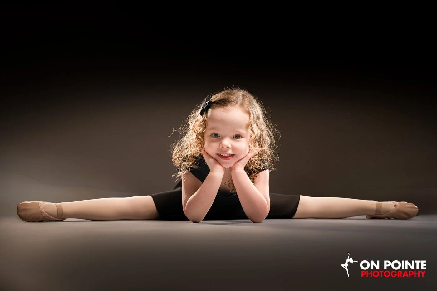 Young girl in ballet pose performing the splits, wearing a black outfit and ballet slippers, smiling at the camera, on a dark background.