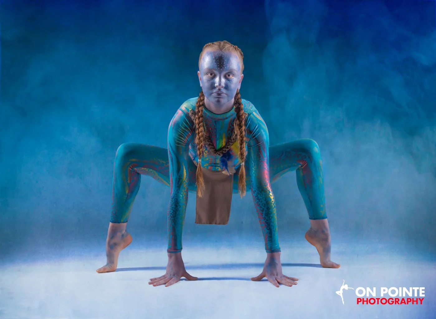 A person in a full-body, colorful costume with face paint and braided hair crouching in a dramatic pose against a blue background.