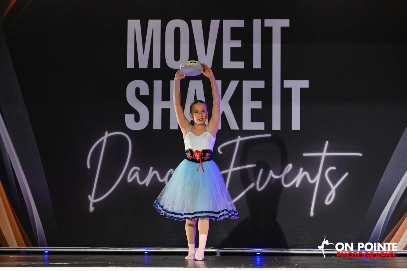 A young dancer in a blue tutu holding a tambourine above her head on stage with 'Move It Shake It Dance Events' in the background.