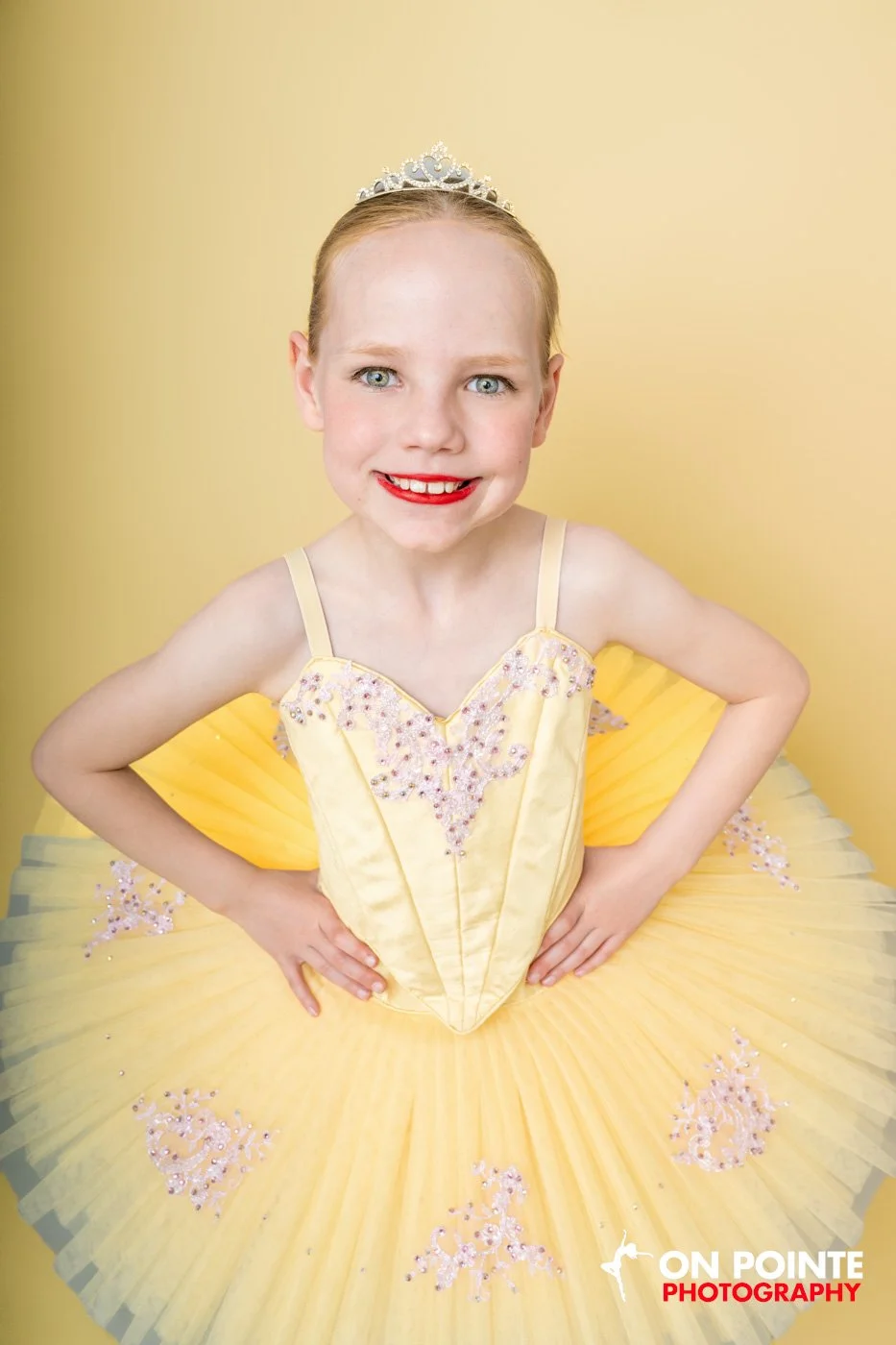 Young girl wearing a yellow tutu and tiara, smiling with hands on hips, against a yellow background.