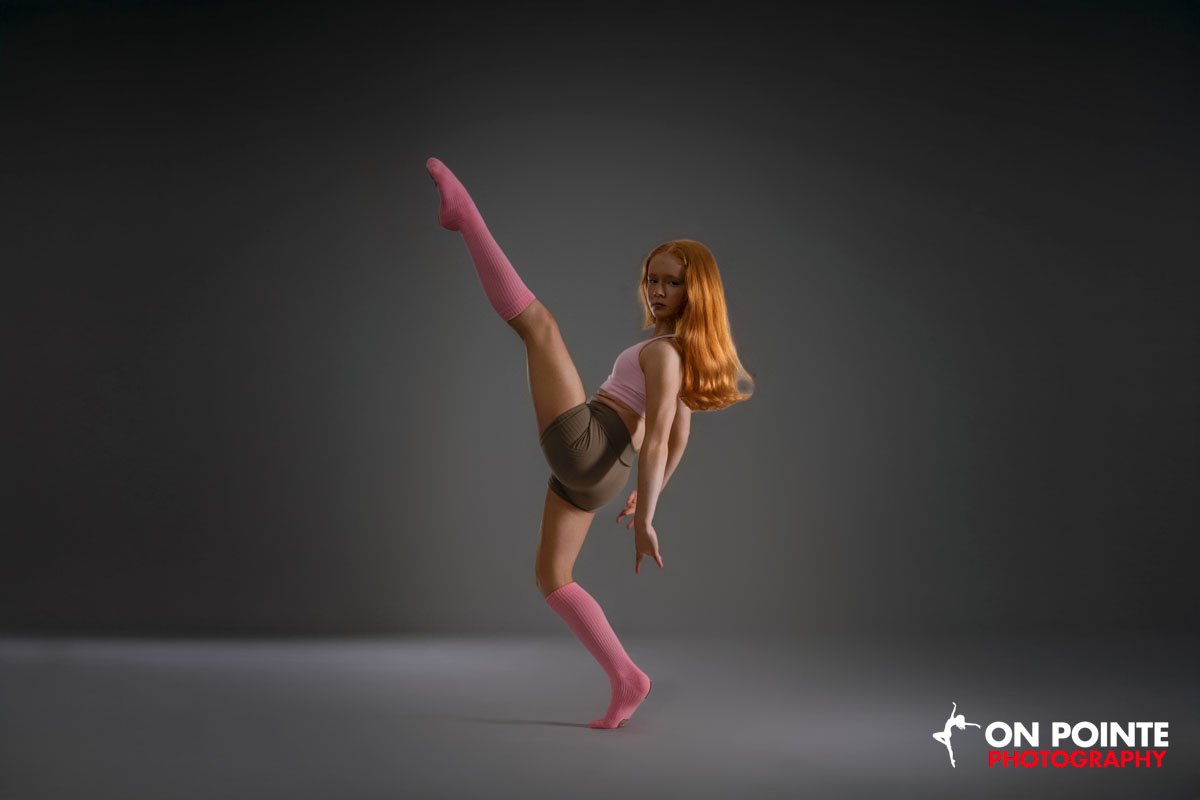 A woman with red hair in a pink tank top, beige shorts, and pink knee-high socks performs a dance pose with one leg raised high, standing on one foot against a plain gray background.