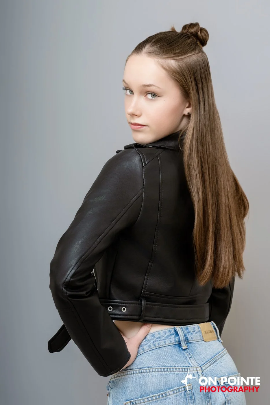 A young woman with long, straight brown hair styled in a half bun, wearing a black leather jacket and light blue jeans, standing against a plain gray background.