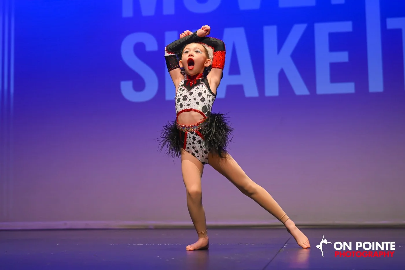 Young girl performing a dance routine on stage, wearing a black, red, and white costume with feathers, with a surprised or excited expression, against a purple background that says 'SHAKE'.