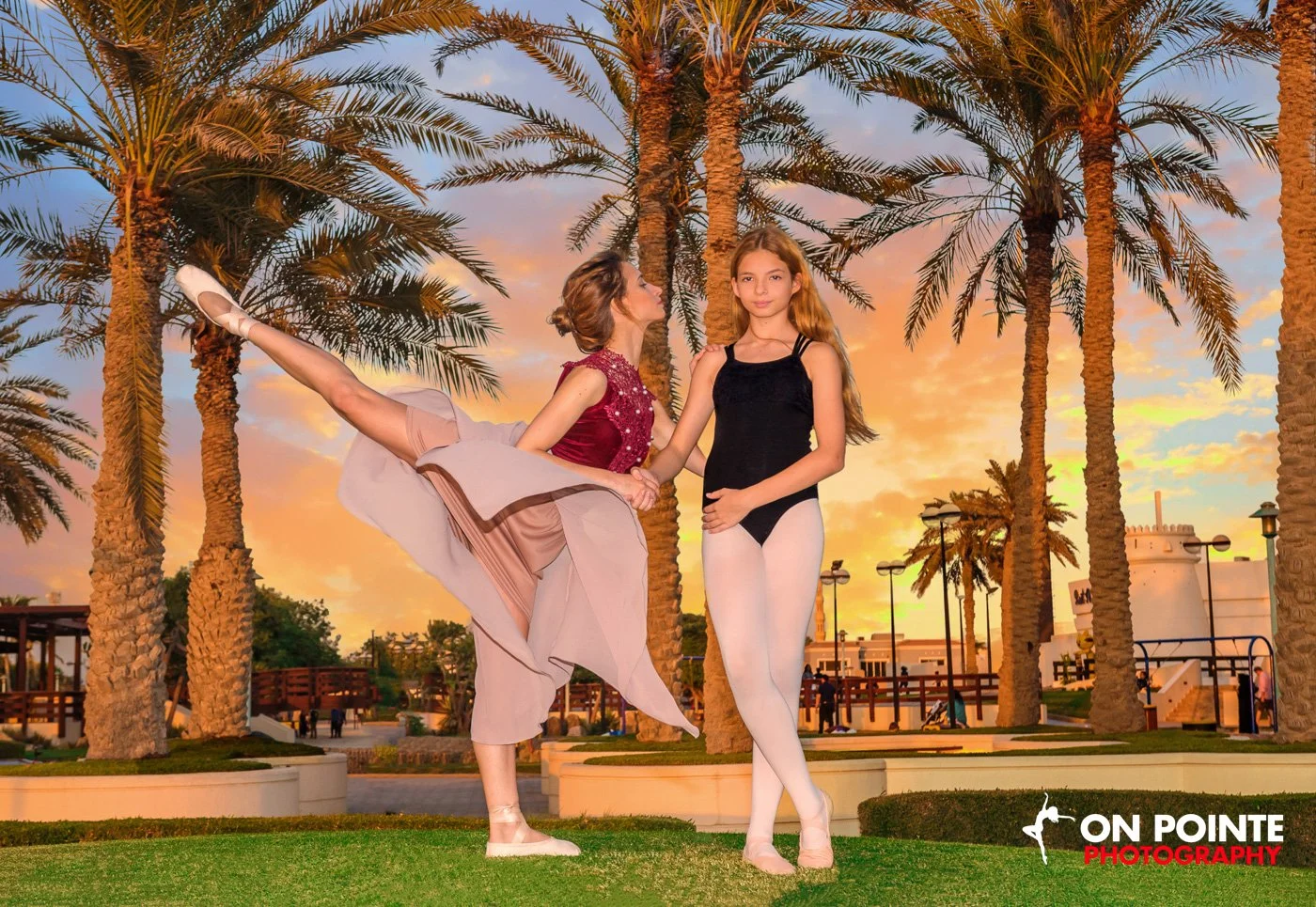 Two ballet dancers posing outdoors near palm trees with sunset sky.