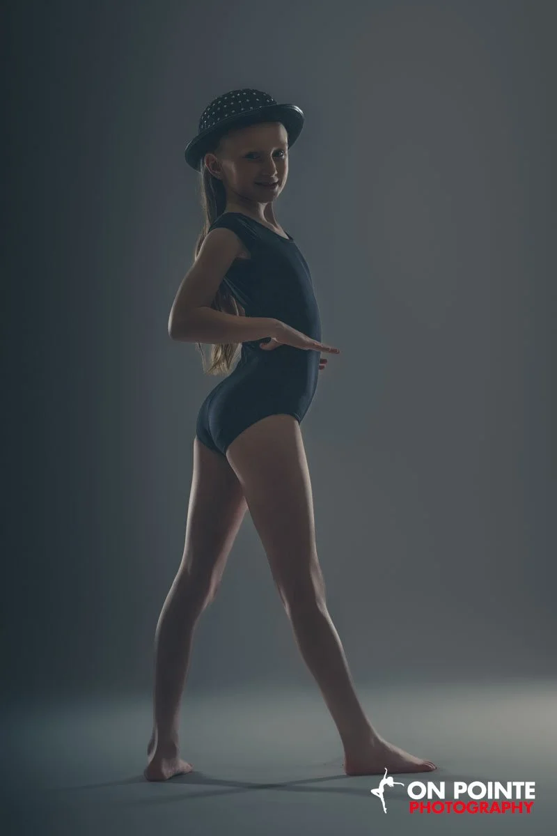 A young girl in a black leotard and a black hat with white polka dots poses with one hand on her hip and the other extended outward, on a gray background. The logo "ON POINTE PHOTOGRAPHY" is at the bottom right corner.