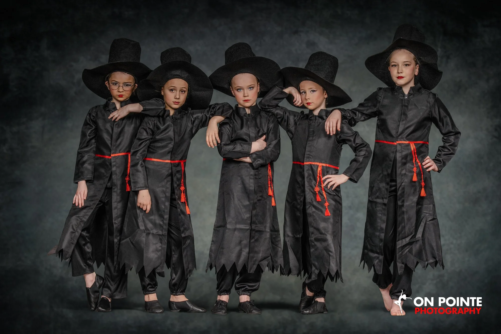 Five young girls dressed as witches in black costumes and large black hats, standing together with serious expressions against a dark background.