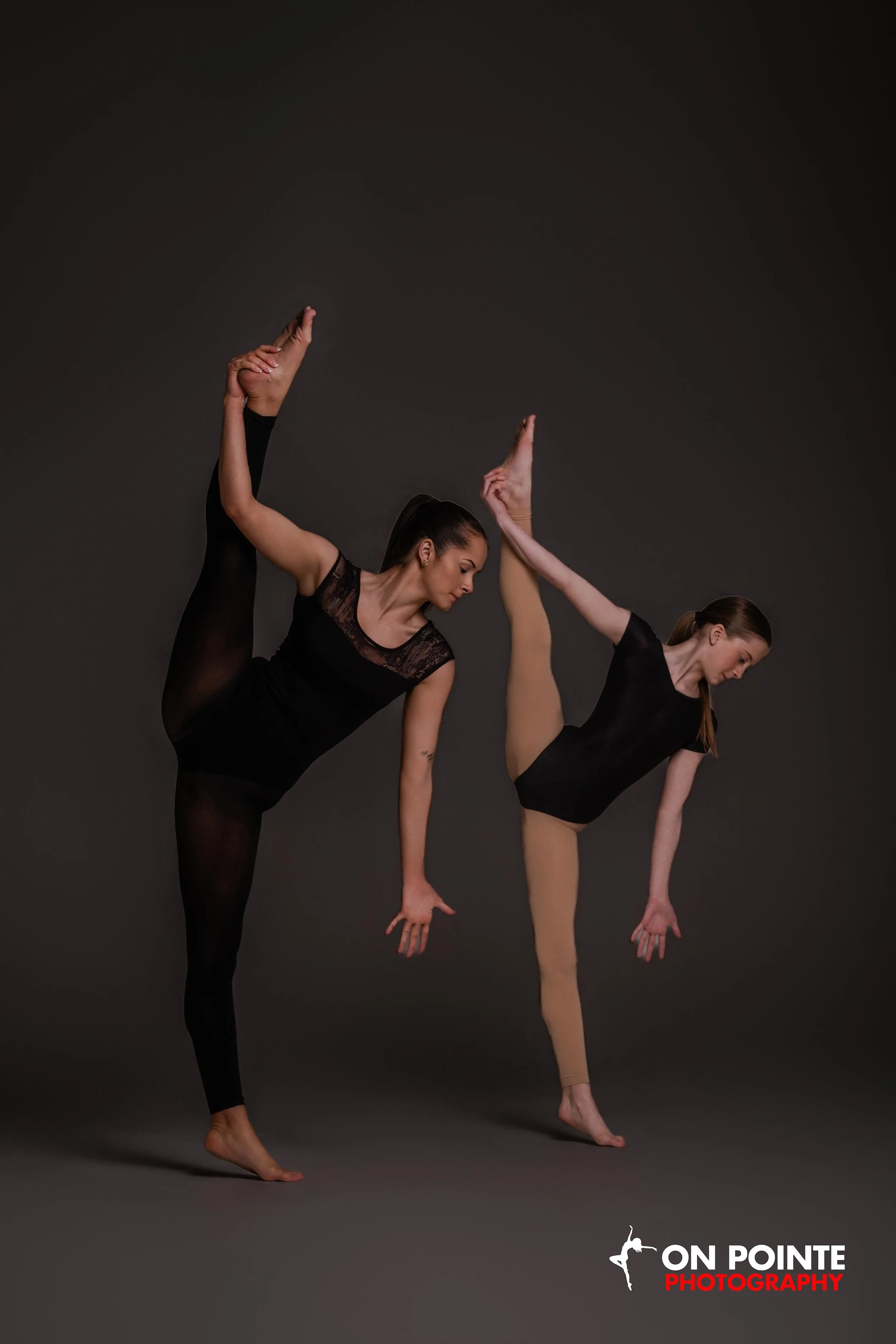 Two women in black dance costumes performing a ballet pose against a dark background, with one woman balancing on one foot and the other raising her leg and arm. "On Pointe Photography" logo appears in the bottom right corner.