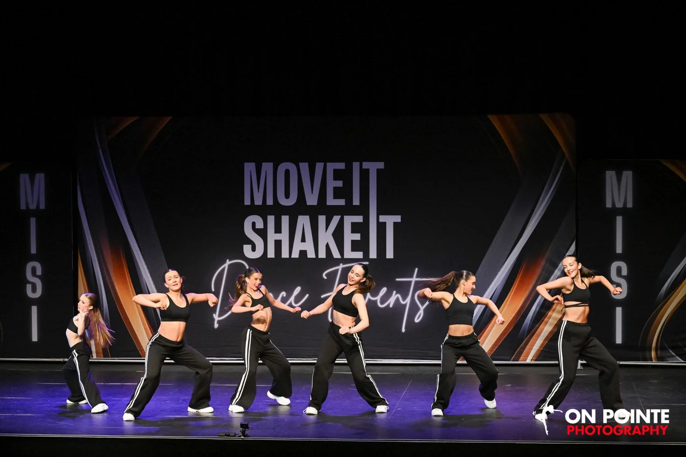Group of dancers performing on stage at 'Move It Shake It' event, wearing black outfits.
