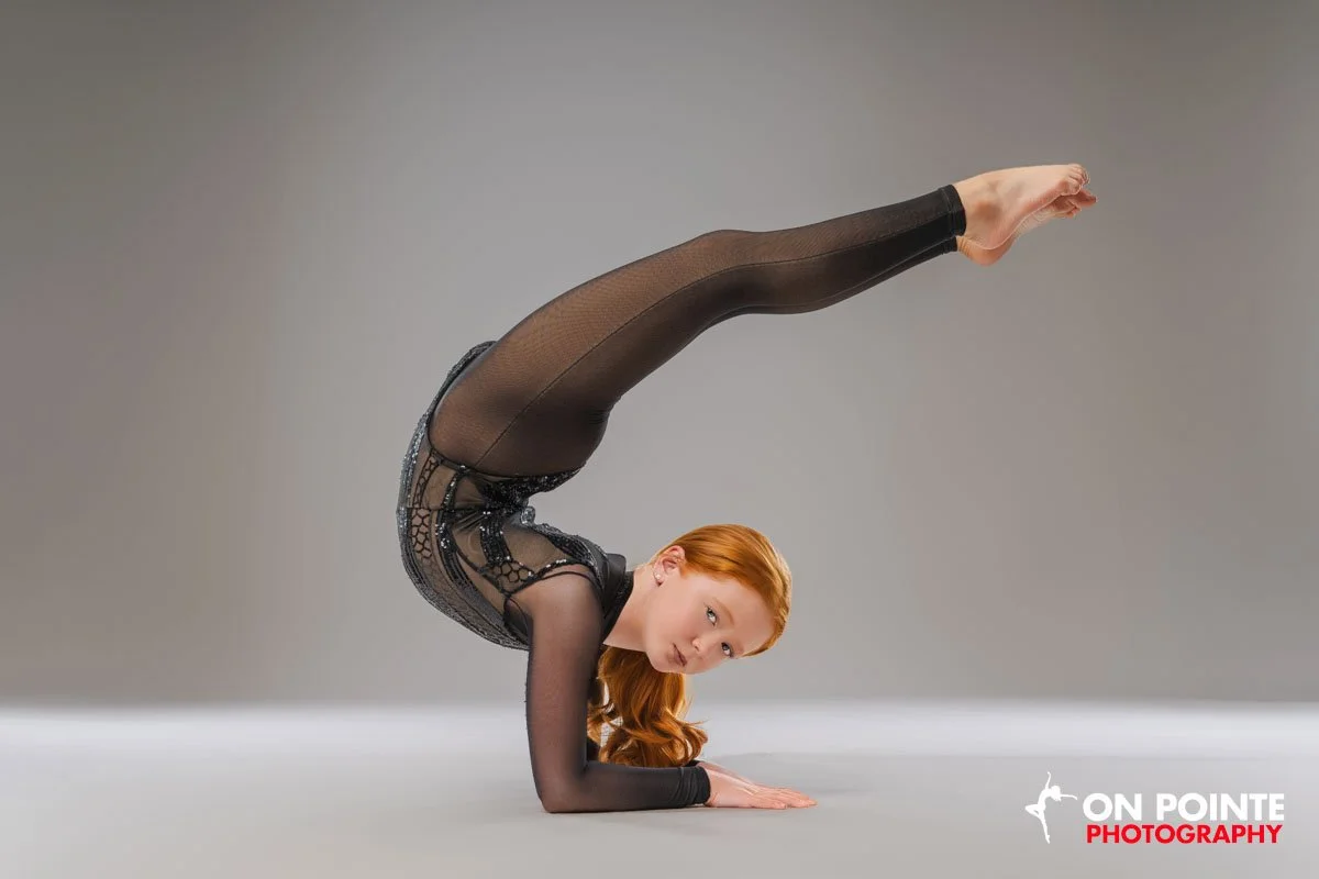 A woman with red hair performing a headstand yoga pose in black sheer athletic wear on a plain gray background.