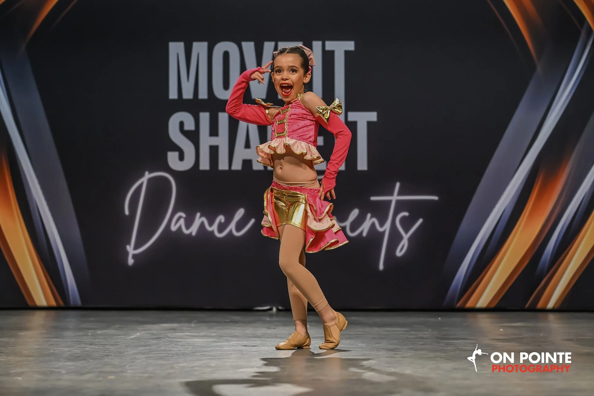Young girl in a pink and gold dance costume performing on stage at a dance competition.