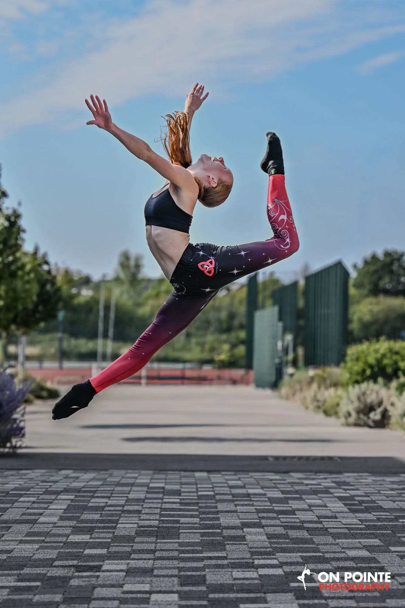 A woman performing a high jump outdoors, with a blue sky and trees in the background. She wears black and pink athletic clothing and has red hair. The ground is paved with a patterned surface, and a logo that says "ON POINTE PHOTOGRAPHY" is in the bo