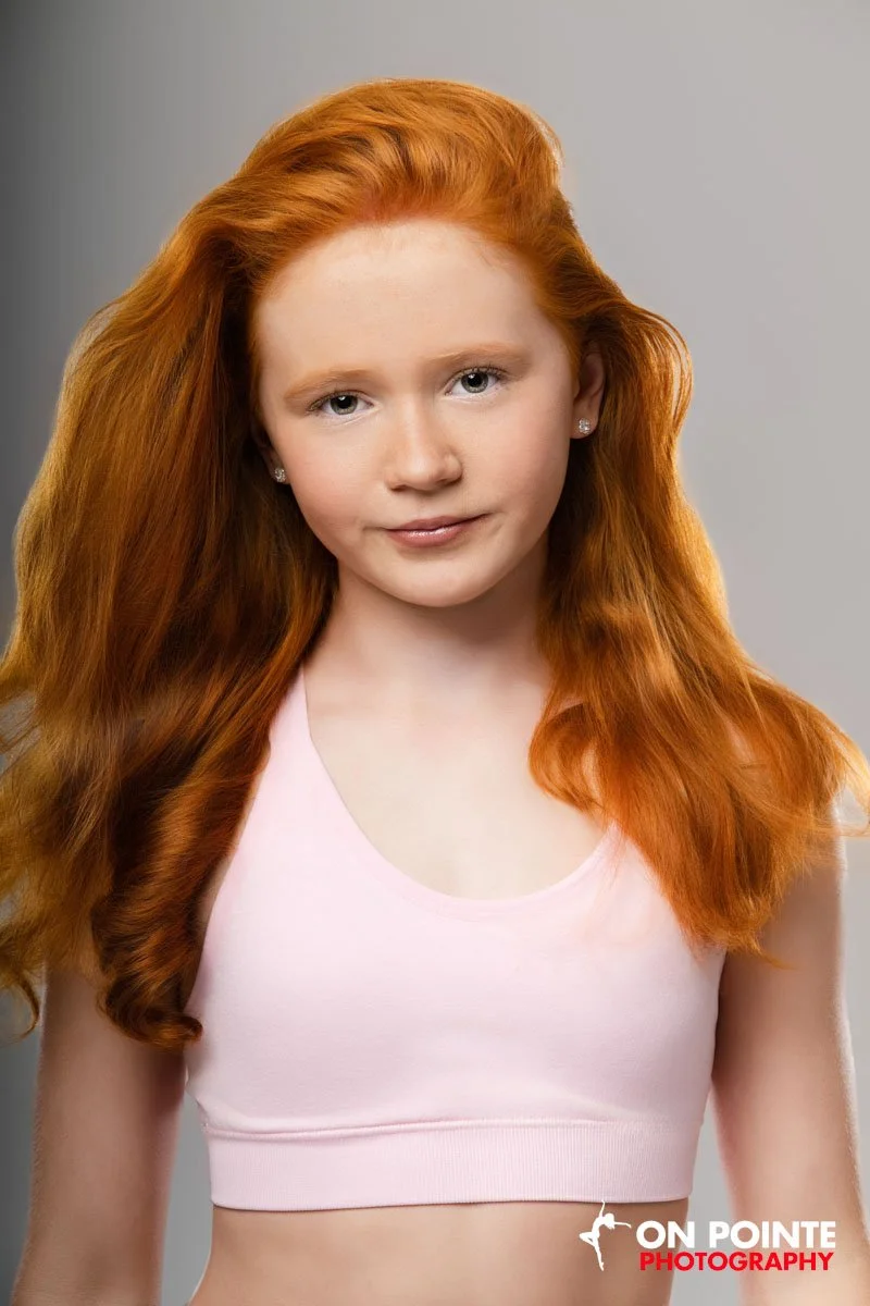 A young woman with long, wavy red hair wearing a light pink sleeveless crop top and small stud earrings, posing against a neutral gray background.