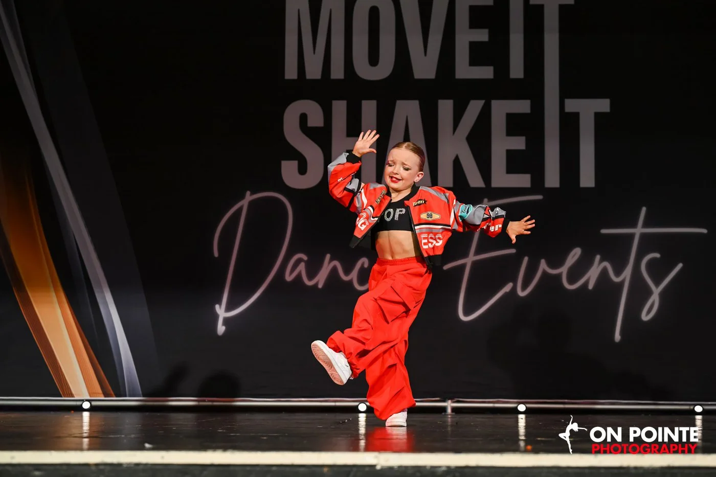Young dancer in red costume performing on stage with "Move It Shake It Dance Events" backdrop.