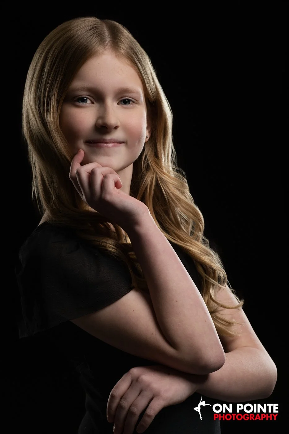 Young girl with long blonde hair wearing a black top, posing with her hand on her chin against a black background.