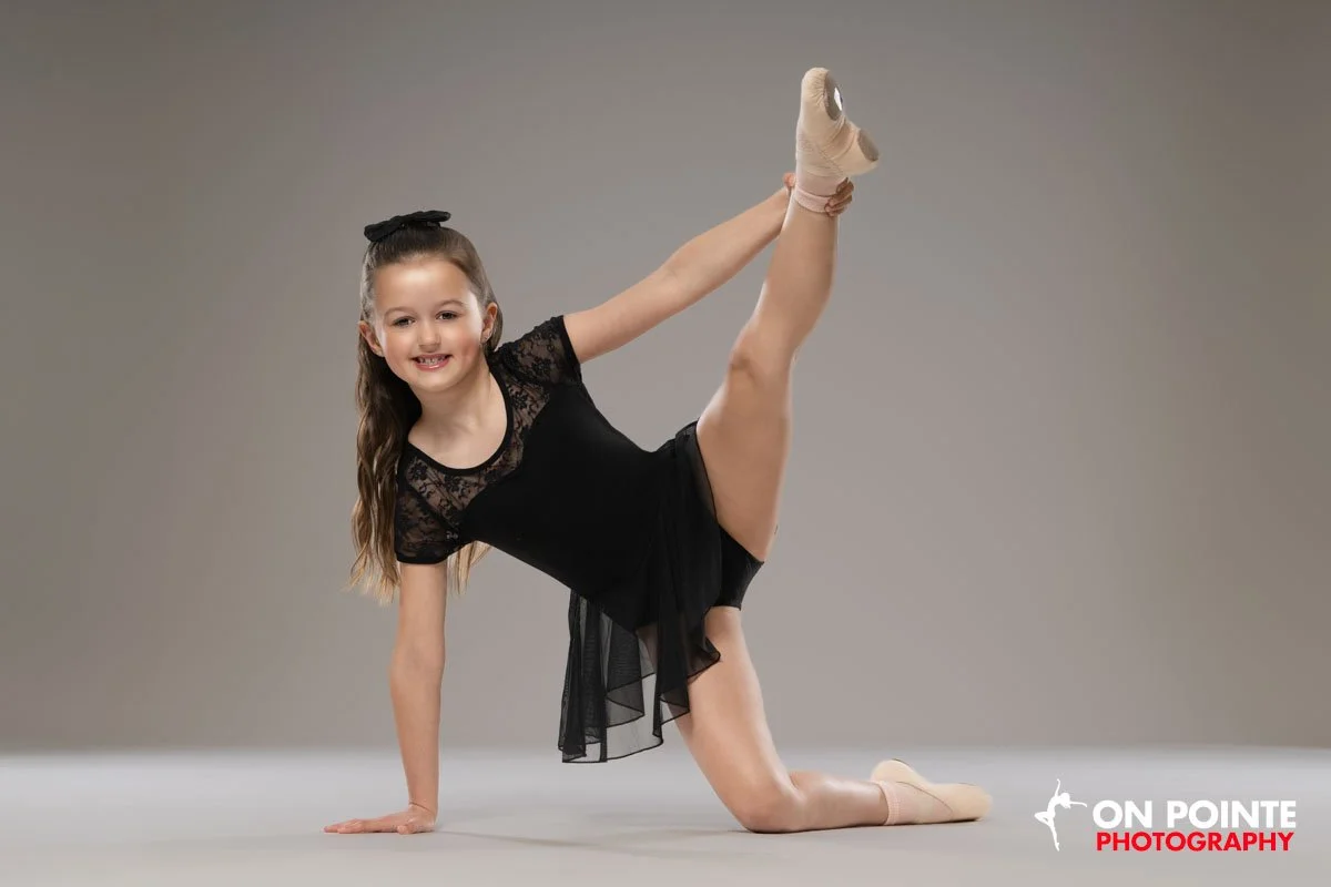 Young girl in a black ballet outfit doing a ballet pose on the floor, smiling at the camera.