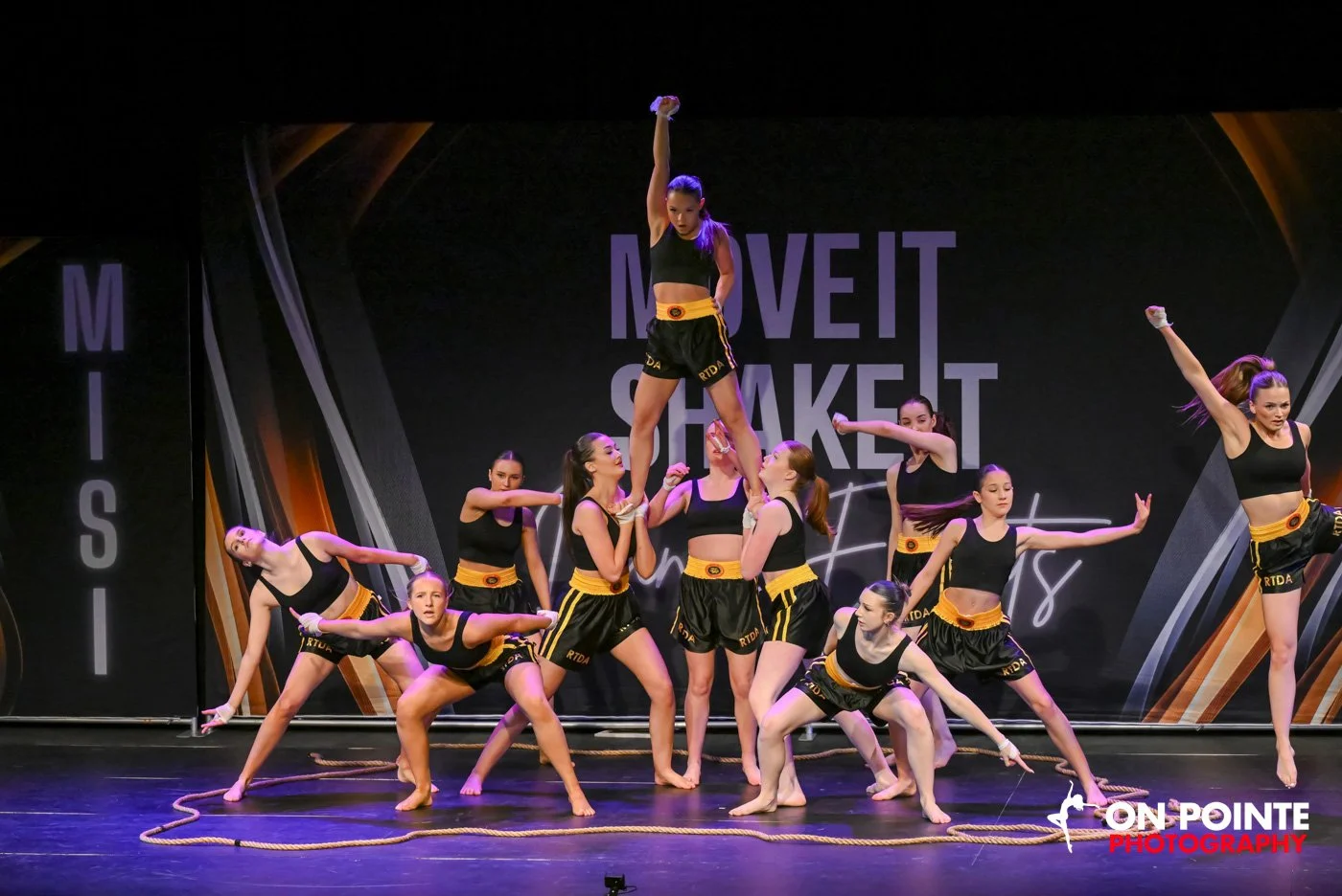 Dance group performing on stage, with one dancer lifted in the air and others in dynamic poses, wearing coordinated black and yellow outfits.