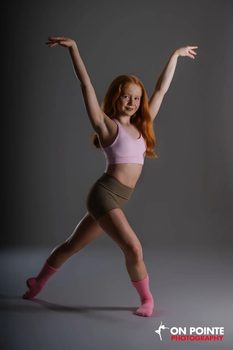 A young girl with red hair wearing pink sports bra, brown shorts, and pink socks poses with arms raised and one leg forward against a plain gray background.