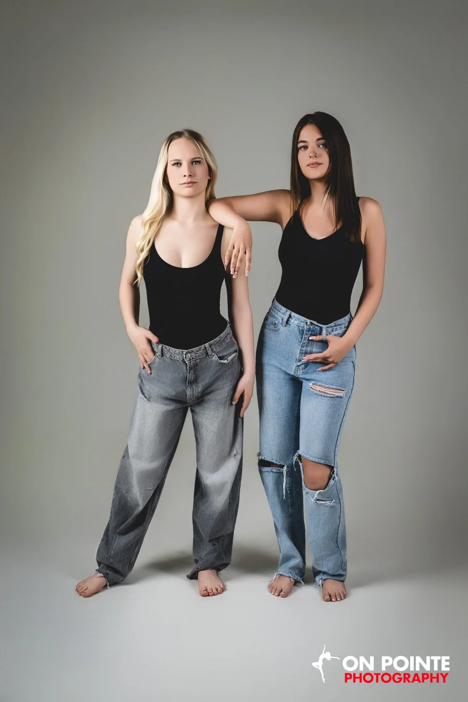 Two young women with different hair colors and styles posing together, wearing black tank tops and jeans, standing against a neutral background, with the logo 'On Pointe Photography' in the bottom right corner.